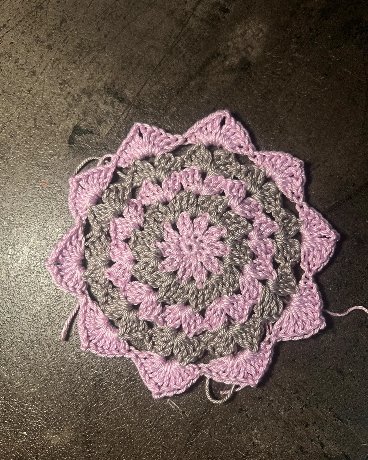Motivo crochet rosa e grigio ancora in lavorazione su superficie scura